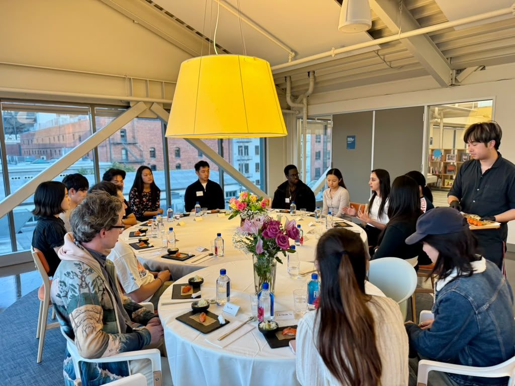 A chef serving nigiri to students seated at a round table by tall windows