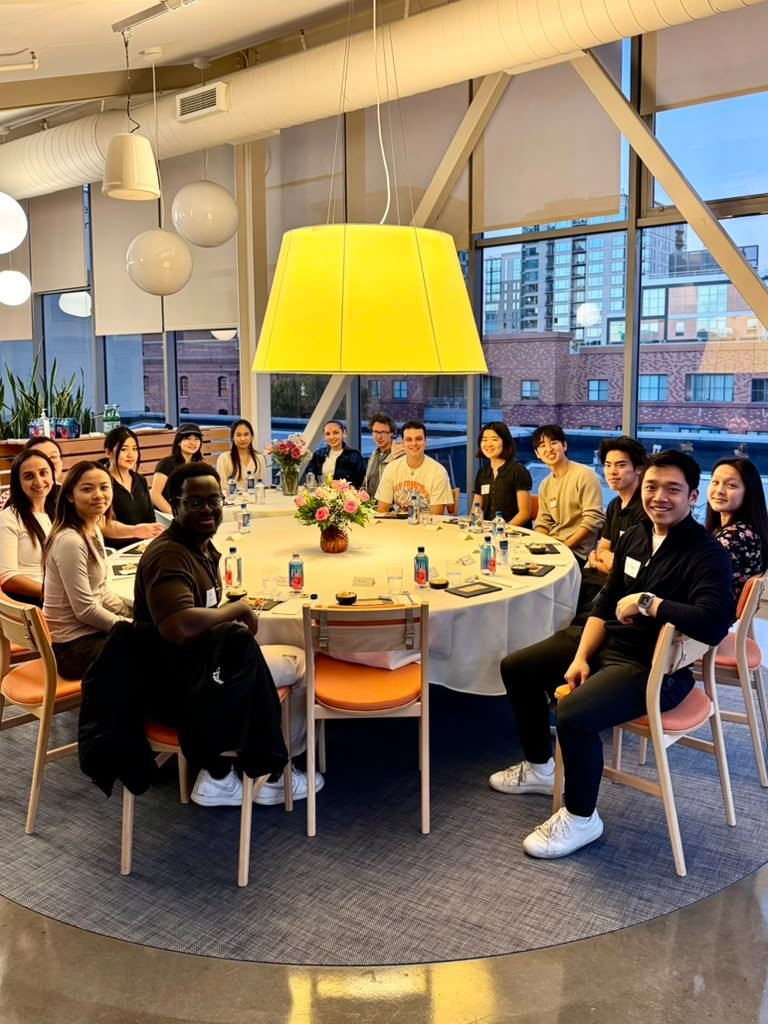 About fifteen students smiling around a round table under a large yellow lamp