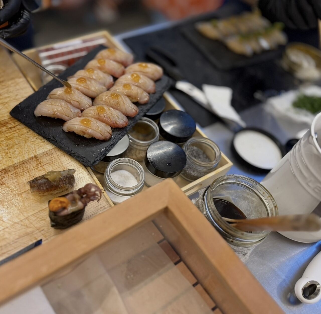 A chef finishing a slate of seared nigiri with a dab of sauce, beside jars of salt and seasonings
